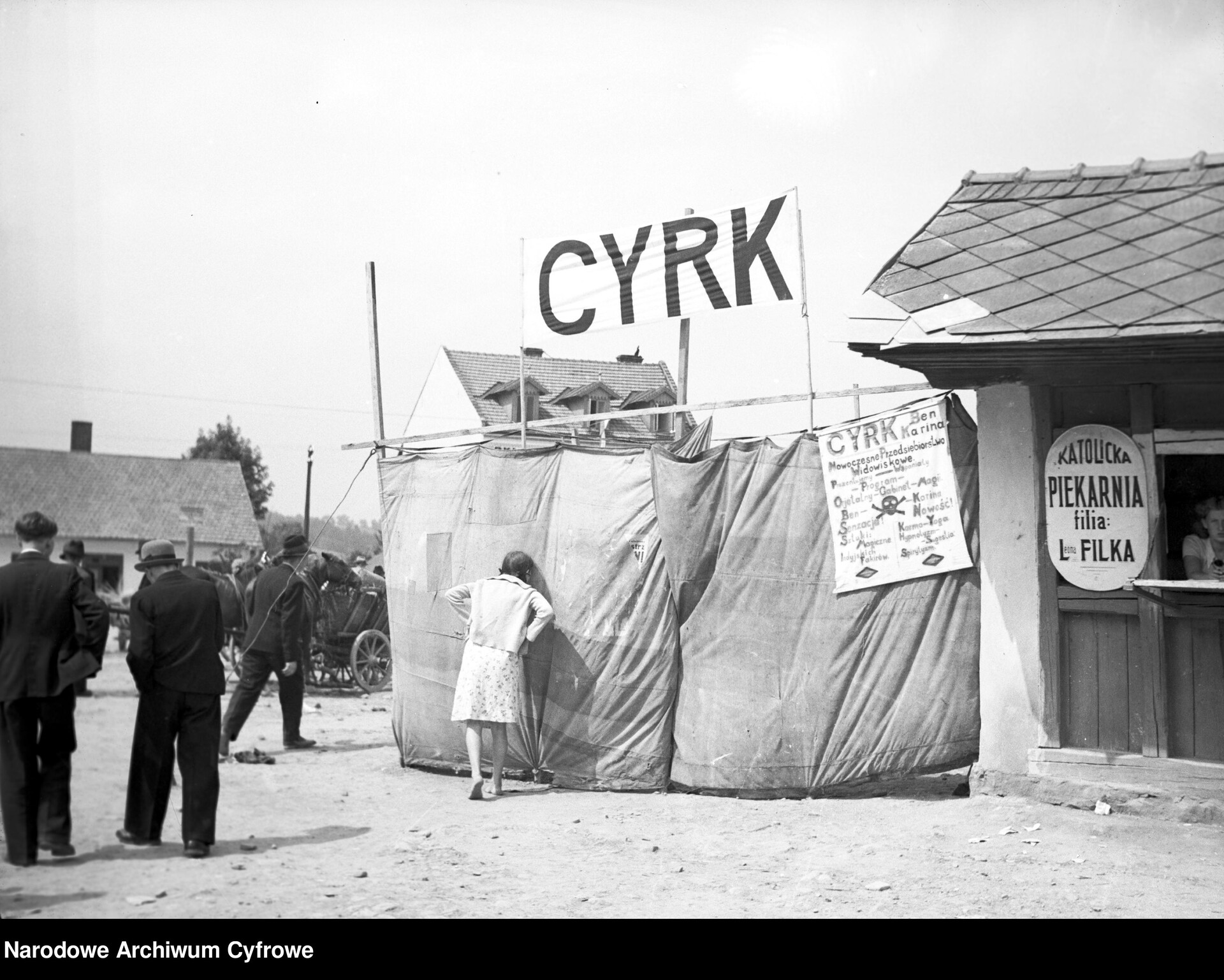  Czarno-białe zdjęcie z lat 30. przedstawia dziewczynkę zaglądającą do cyrku przez dziurę ogrodzenia wykonanego z materiału. Z boku widoczna jest plansza reklamująca cyrk oraz szyld „katolickiej piekarni”. 