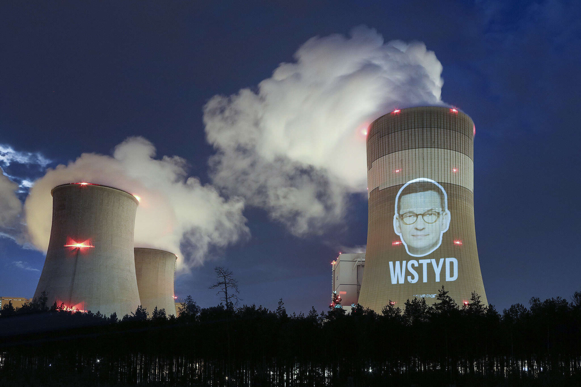  Zdjęcie przedstawia dymiące kominy elektrowni Bełchatów. Na jednym z kominów za pomocą projektora została wyświetlona twarz Mateusza Morawieckiego, a pod nią napis „wstyd” zapisany dużymi literami. 