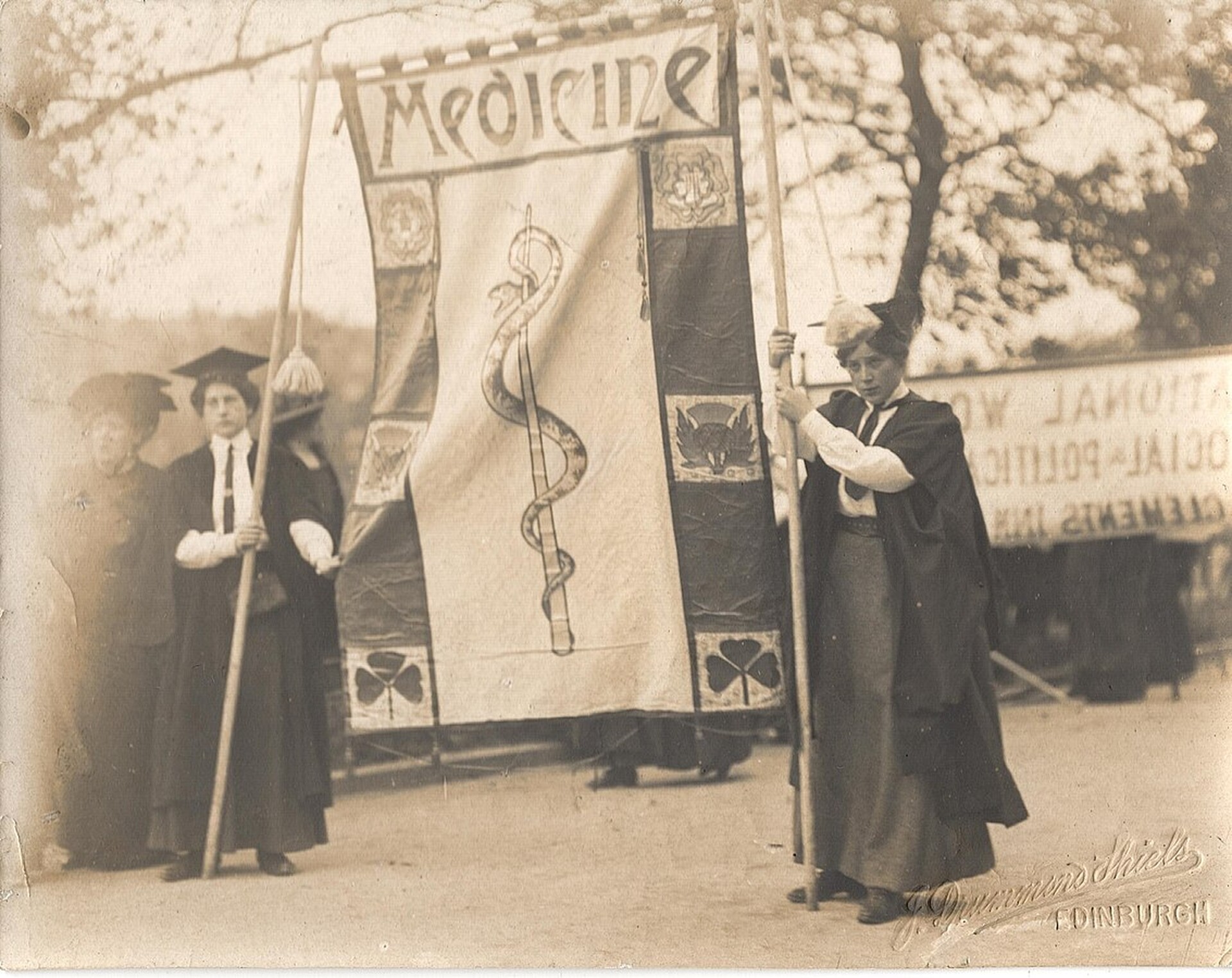  Czarno-białe zdjęcie z 1908 roku. Przedstawia dwie młode sufrażystki trzymające banner. Banner jest wysoki na około dwa metry i wykonany z materiału. Na górze banneru znajduje się napis „Medicine”, a pod nim laska Eskulapa 