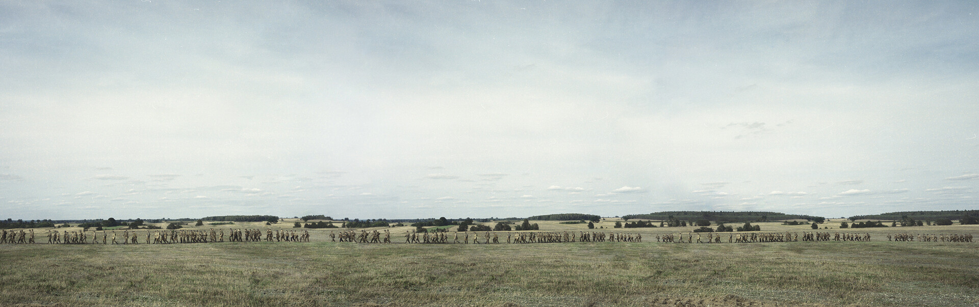  Fotografia Agnieszka Rayss Przemarsz z cyklu A więc wojna. Długie, panoramiczne zdjęcie przedstawiające przemarsz osób w wojskowych mundurach przez pole. 